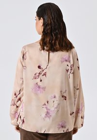 Blusa beige chiaro con stampa floreale in tonalità di rosa e bordeaux. Texture liscia, maniche lunghe, dettaglio sul retro a chiave e vestibilità morbida.