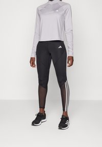 Vaaleanharmaa pitkähihainen paita, jossa on korkea kaulus, yhdistettynä mustiin leggingseihin, joissa on verkkopaneelit ja kolme valkoista raitaa sivuilla. Mustat lenkkarit.