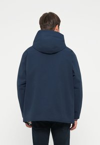 Giacca a vento blu navy con colletto alto e grande cappuccio. Caratterizzata da una vestibilità rilassata e un tessuto liscio e resistente all'acqua.