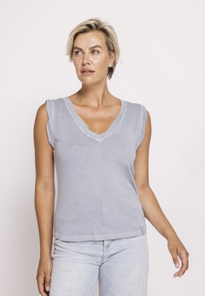 Blonde Frau trägt ärmelloses hellblaues V-Ausschnitt-Top und Jeans mit hellem Waschmittel, schaut leicht nach links vor einem schlichten Hintergrund.