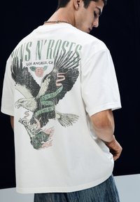 Hvid oversized t-shirt med et ørnerelief på ryggen, med teksten "Guns N' Roses," og blødt tekstureret bomuldsstof.