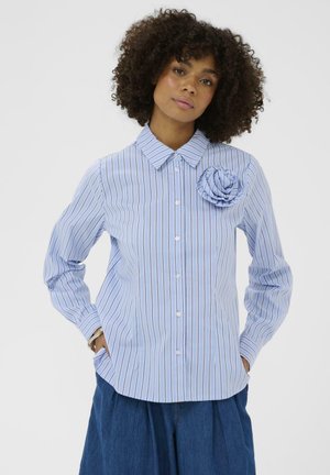 Göndör hajú nő kék csíkos ingben, gombokkal, textilvirág díszítéssel, kezét a zsebében tartva, egyszerű háttér előtt áll.