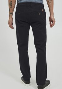 Pantaloni negri din bumbac cu croială dreaptă, prevăzuți cu buzunare la spate cu închidere prin nasture și o textură simplă. Purtați cu adidași negri.