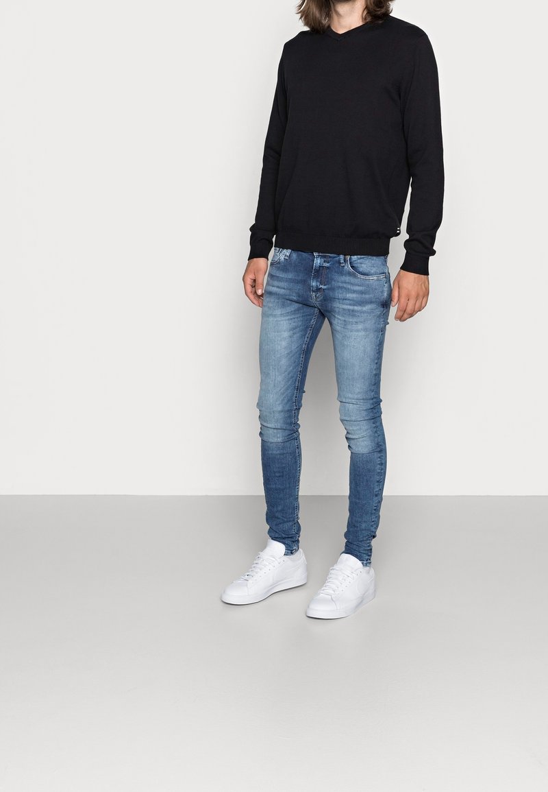 Pull noir à manches longues avec col en V, jean skinny bleu clair, et baskets blanches. La tenue est simple avec une silhouette décontractée et ajustée.