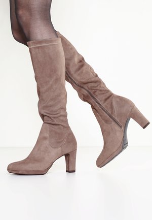 Kvinde iført taupe ruskind knæhøje støvler med blokhæle og sidelåse, kombineret med gennemsigtige sorte strømpebukser.