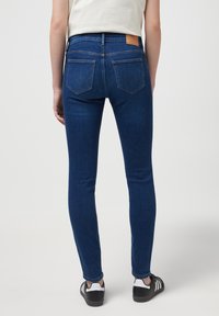 Un jean en denim bleu ajusté présente une taille mi-haute, des poches arrière avec des coutures en biais, et une étiquette en cuir beige à la ceinture arrière.