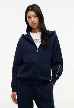 Ung kvinde iført en marineblå lynlåshoodie og matchende bukser med en hvid Tommy Hilfiger t-shirt synlig under.