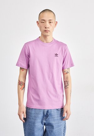 Camiseta de algodón de color púrpura claro con manga corta y escote redondo; presenta un pequeño logo negro de Adidas en el lado izquierdo del pecho. Combinada con jeans azules.