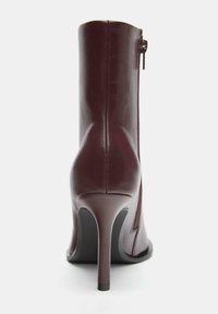Bourgognefarvet læderankelstøvle med høj stilethæhæl, sidelås, glat tekstur og slankt design. Bagfra fremhæver dens kurver.
