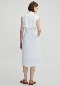Witte jurk met geborduurde kapmouwen, een getailleerde taille en een rok tot de knie. Casual ontwerp, gladde stof met een lichte glans. Bruine sandalen.