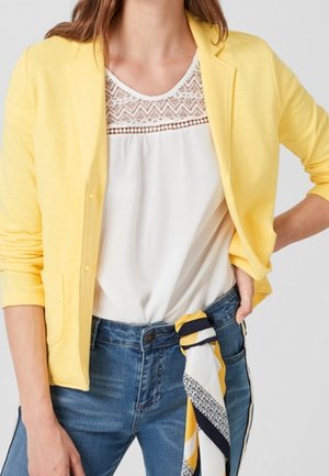 Vrouw draagt een gele blazer over een wit kanten topje, blauwe spijkerbroek met een geel-wit-zwart patroon sjaal vastgeknoopt aan de taille.