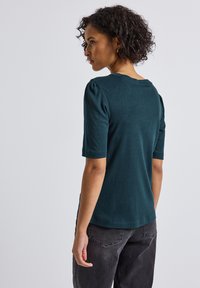Haut turquoise à manches courtes avec un col arrondi et de légères fronces aux épaules, associé à un jeans gris foncé, vu de dos.