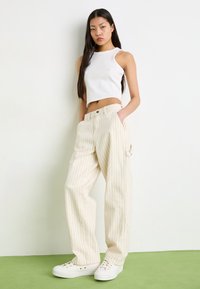 Lightweight, cropped white tank top paired with wide-legged, cream pants featuring red vertical stripes and side pockets. White sneakers visible.