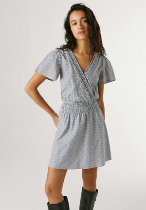 Femme portant une robe portefeuille à manches courtes à rayures noires et blanches, avec une taille froncée, debout, une main sur la hanche et des bottes noires.