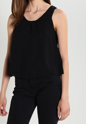 Mujer con camiseta negra sin mangas con cuello redondo y pantalones negros, de pie contra un fondo claro y liso.