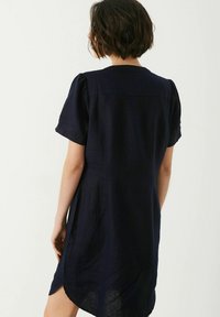 Nainen, jolla on lyhyet tummat hiukset, pukeutuneena lyhythihaiseen laivastonsiniseen mekkoon, seisoo selkä katsojaan päin yksinkertaista taustaa vasten.