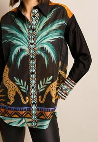 Chemise noire boutonnée avec un motif de palmier et d'imprimé léopard en turquoise et orange, avec des motifs géométriques sur les poignets et la patte de boutonnage.