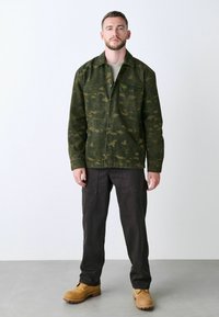 Veste en camouflage vert avec des boutons et des poches latérales, associée à un pantalon cargo foncé et des bottes de travail beige. Tissu lisse et texturé.