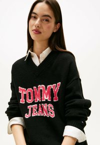 Maglione nero a scollo a v con la scritta "TOMMY JEANS" in grassetto rosa, tessuto testurizzato e una camicia bianca di sotto con maniche arrotolate.