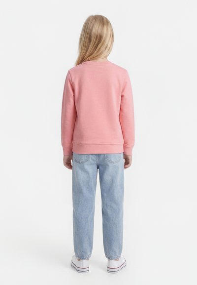 Παιδί με μακριά ξανθά μαλλιά στέκεται με την πλάτη γυρισμένη, φοράει ροζ φούτερ, ανοιχτό μπλε τζιν και λευκά αθλητικά παπούτσια, σε απλό φόντο.