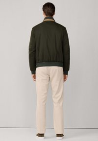 Uomo in giacca bomber verde scuro, pantaloni beige e scarpe marroni si trova di fronte a un muro grigio chiaro semplice.