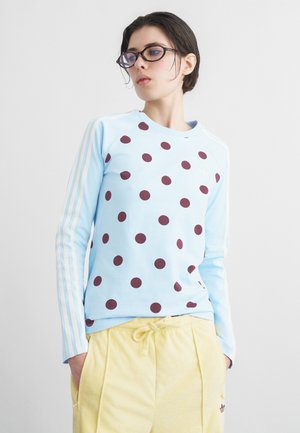 Mladý človek s krátkymi tmavými vlasmi a okuliarmi, ktorý má na sebe svetlomodrú mikinu s bodkami a bledozelené nohavice so šnúrkou.