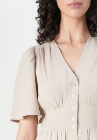 Abito beige chiaro con maniche a sbuffo corte, scollo a V e sei bottoni decorativi sulla parte anteriore. Tessuto con texture e dettaglio in vita arricciato.
