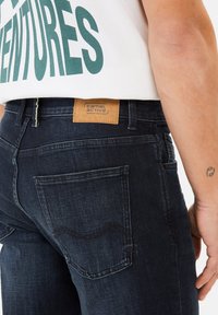 Donkerdenim jeans hebben een gladde textuur, rechte snit en een tan lederen merkaffiche met het label "Camel Active" op de achterkant van de tailleband.