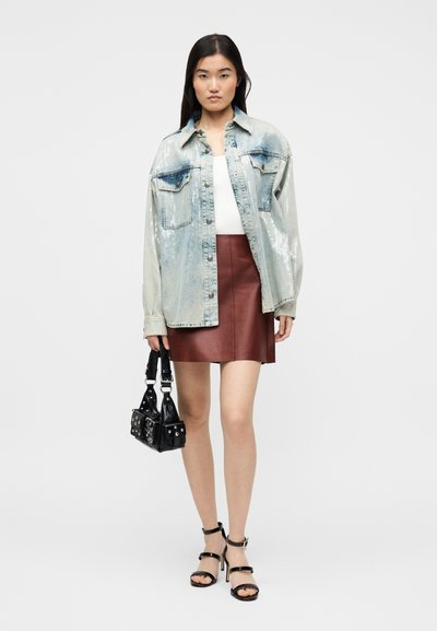 Woman wearing a sequined denim jacket, white top, brown leather skirt, black high-heeled sandals, and holding a black studded handbag.