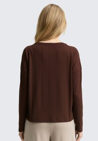 Vrouw met lang blond haar in een geribbelde donkerbruine lange mouwtop en lichtbeige broek, van achteren gezien tegen een eenvoudige achtergrond.