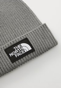Szürke kötött sapka bordázott textúrával, fekete téglalap alakú folttal, amelyen fehér színű "THE NORTH FACE" logó található.