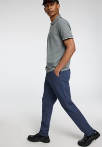 Grijze polo met witte accenten, gecombineerd met donkerblauwe broeken en zwarte schoenen. Het model staat in profiel met de handen in de zakken.