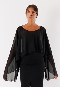 Cappa nera in chiffon con maniche ampie, design a strati e scollatura arrotondata, indossata sopra una gonna nera aderente con uno spacco.