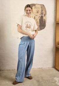 Femme souriante ajustant un t-shirt beige ample avec un imprimé crabe, portant un pantalon large bleu clair et des chaussures marron, debout dans un espace intérieur neutre.