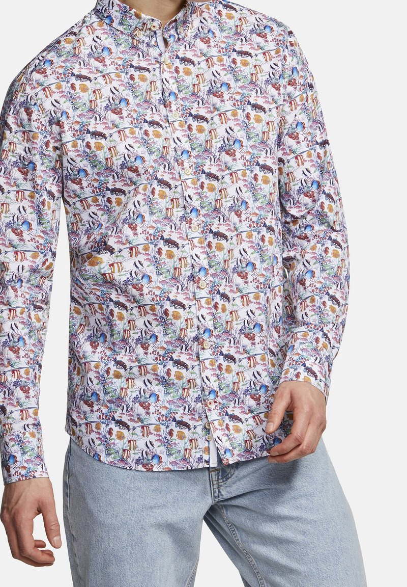 Homme portant une chemise à manches longues boutonnée avec un motif coloré de poissons et de corail, associée à un jean bleu clair.