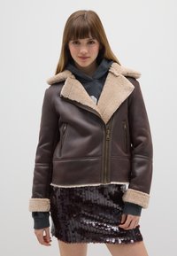 Giacca di pelle marrone con colletto in shearling beige, chiusura frontale con cerniera, due tasche laterali e maniche con polsini. Indossata sopra una felpa con cappuccio grigia.