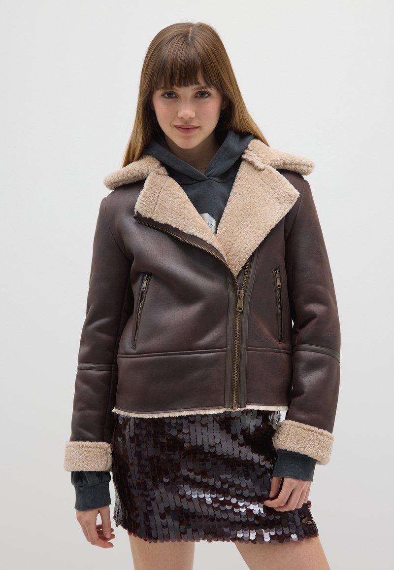 Giacca di pelle marrone con colletto in shearling beige, chiusura frontale con cerniera, due tasche laterali e maniche con polsini. Indossata sopra una felpa con cappuccio grigia.