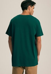 Zielona bawełniana koszulka z luźnym krojem, krótkimi rękawami i okrągłym dekoltem. Gładka faktura z czystym wykończeniem, noszona z beżowymi spodniami.