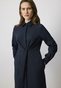 Rochie bleumarin cu dungi, cu guler butonat, mâneci lungi și detaliu cu nod la talie. Confecționată dintr-un material neted, cu o croială ajustată.