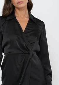 Abito avvolgente in satin nero con maniche lunghe, colletto e dettagli plissettati in vita, caratterizzato da una superficie liscia e una caduta elegante.