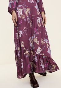 Floral Maxi-Kleid mit lila Hintergrund, ausgestattet mit botanischen Designs in Hellblau und Gold. Rüschensaum und lange Ärmel, weicher Stoff.