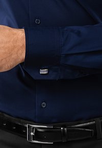 Chemise bleu marine avec une texture lisse, poignets boutonnés et boutons de manchette en argent. Ceinture en cuir noir avec une boucle brillante.