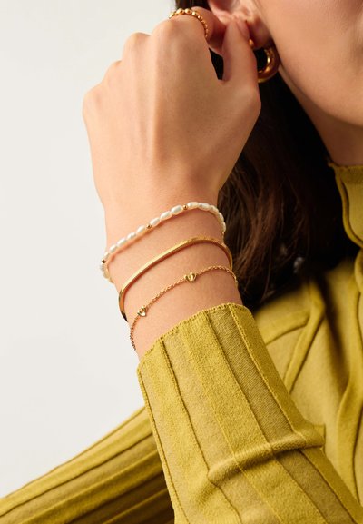 Χέρι με τρία βραχιόλια: λευκή πέρλα με χρυσά χάντρες, χρυσή επίπεδη αλυσίδα και λεπτή χρυσή αλυσίδα με μικρά καρδιές, φορώντας μουσταρδί πλεκτό πουλόβερ με ραβδώσεις.