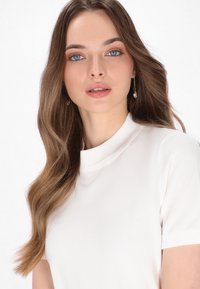 Maglietta bianca a maniche corte con collo alto e texture morbida. La modella ha i capelli lunghi e mossi e indossa orecchini d'oro con accenti di perle.
