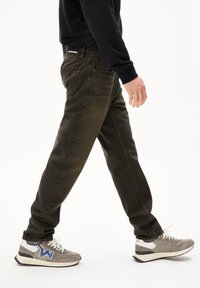 Jeans de mezclilla verde oscuro con corte recto, que presentan bolsillos traseros y detalles de costura sutiles, combinados con zapatillas grises y una camiseta negra.