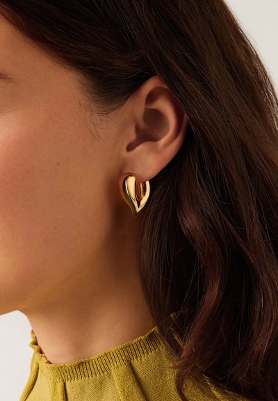 Κοντινό πλάνο στο αυτί μιας γυναίκας που φοράει λαμπερό, χρυσό, αφηρημένο σκουλαρίκι σε σχήμα καρδιάς, με σκούρα μαλλιά και ορατό γιακά ζιβάγκο μουσταρδί χρώματος.