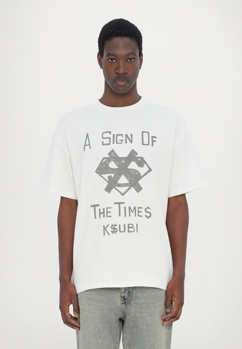 White cotton t-shirt featuring a graphic design in gray with text "A Sign Of The Times K$UB!" and a dollar sign motif. Relaxed fit.