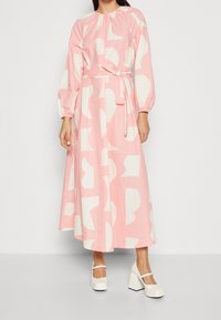 Růžové bavlněné šaty s bílými abstraktními tvary, dlouhými rukávy, nařaseným výstřihem a opaskem na zavazování v pase. Nosené s bílými chunky podpatky.