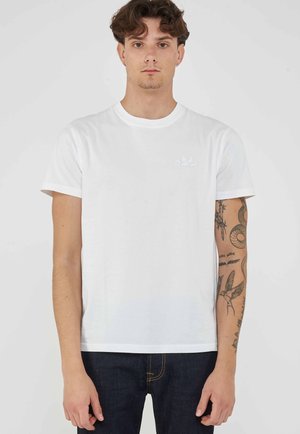 Jonge man met krullend haar, gekleed in een eenvoudig wit T-shirt en donkere spijkerbroek, toont gedetailleerde tatoeages op zijn rechter onderarm tegen een lichte achtergrond.