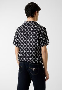 Chemise noire à manches courtes avec un motif géométrique blanc, col, et coupe décontractée. Associée à un jean foncé avec des surpiqûres jaunes.